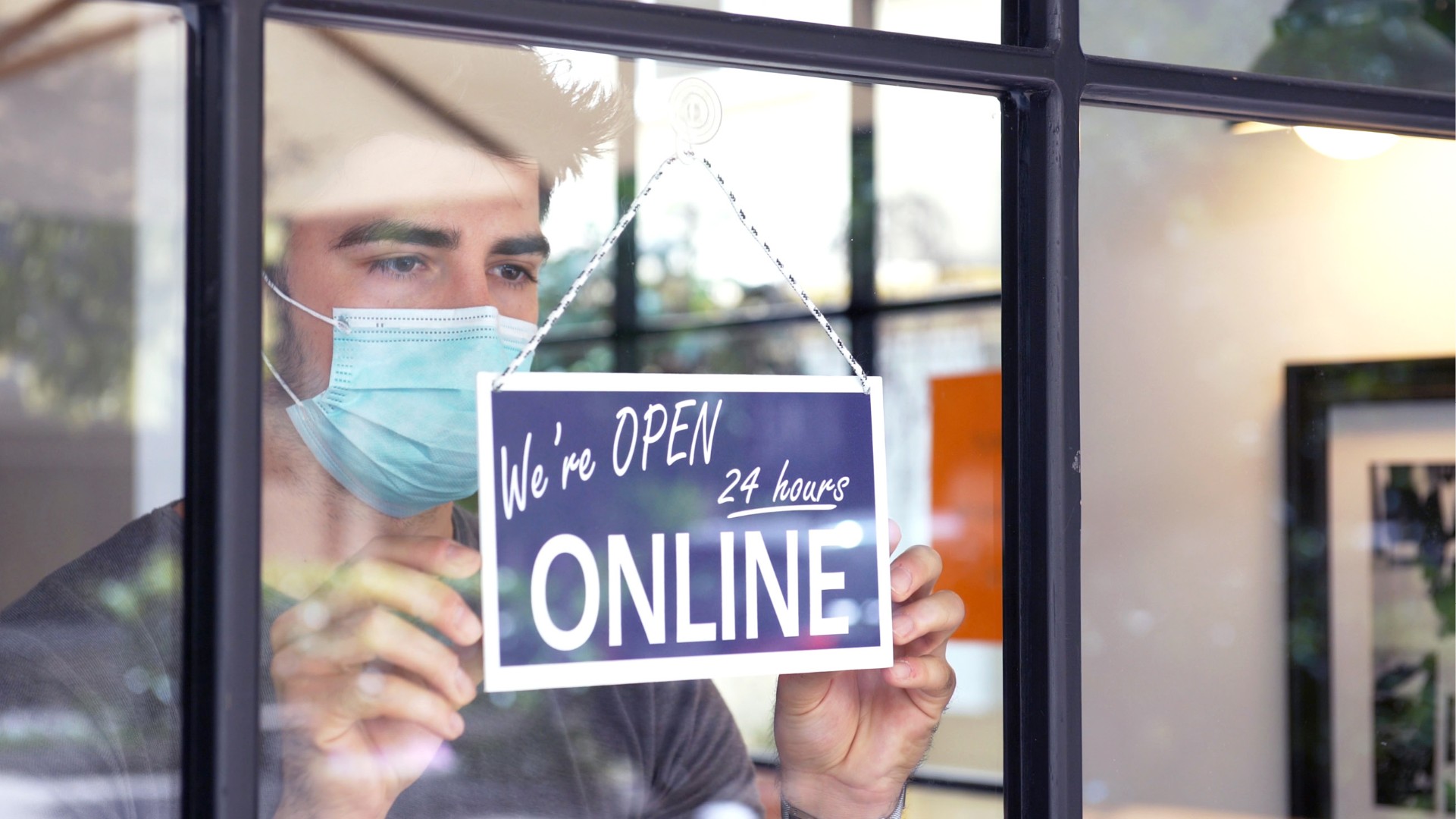 Young man with mask hangs sign on shop door: open 24 hours online